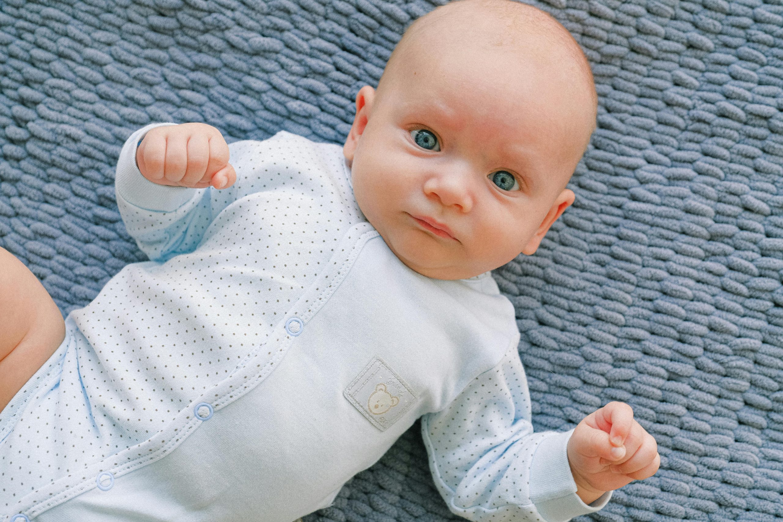 Cute baby in polka dot onesie lying on a textured blue blanket, looking up with a calm expression.